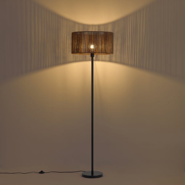 Eingeschaltete Stehleuchte mit zylindrischem Geflechtschirm, die ein dekoratives Streifenmuster aus Licht und Schatten an die Wand wirft.