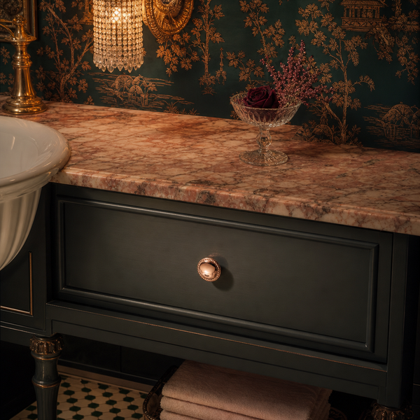 Badezimmer-Unterschrank mit dunkelgrüner Front, kupferfarbenem Knopf und rosa Marmorplatte vor gemusterter Tapete.