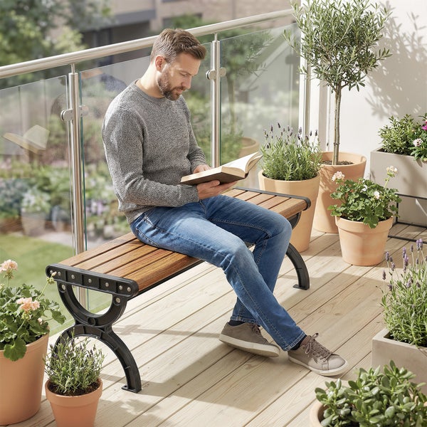 Mann liest ein Buch auf einer Gartenbank mit Holzlatten und Metallgestell auf einem Balkon mit Topfpflanzen und Glasgeländer.