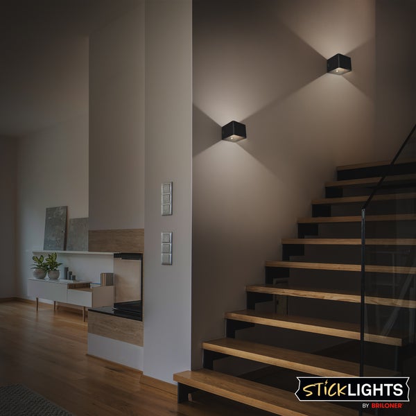 Zwei schwarze, quadratische LED-Wandleuchten an einer Wand neben einer Holztreppe, die Licht nach oben und unten werfen. Sticklights by Briloner Logo.