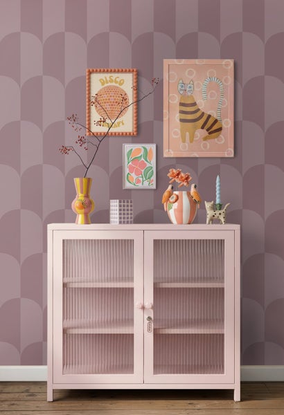 Vliestapete mit Bogenmuster in Mauve hinter einer rosa Kommode mit Glastüren und farbenfroher Dekoration.