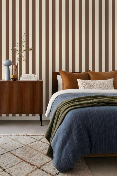 Einrichtung eines Schlafzimmers mit gestreifter Tapete, einem Holzsideboard, einem Bett mit blauer Tagesdecke und einem strukturierten Teppich.