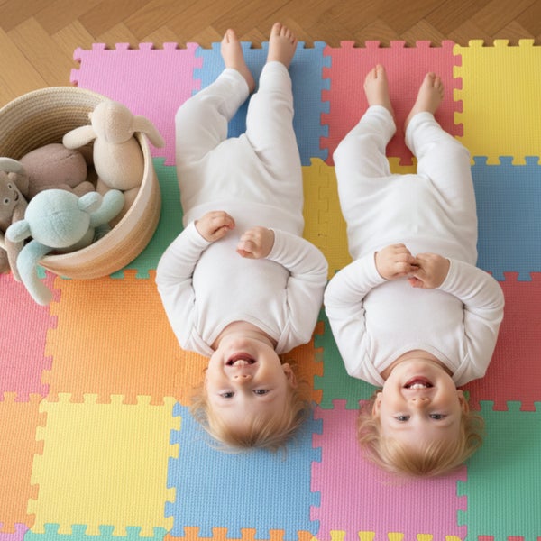 Zwei Kleinkinder liegen auf einer bunten Puzzlematte aus Schaumstoff neben einem Korb mit Kuscheltieren auf einem Holzfußboden.