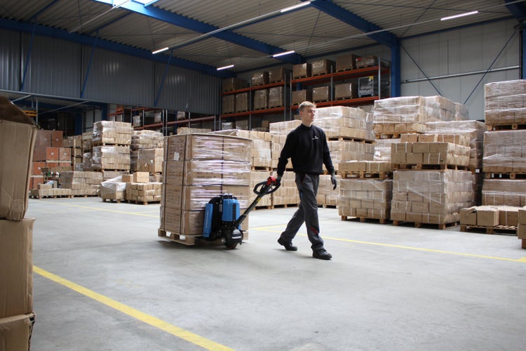 Eine Person bedient einen Elektro-Niederhubwagen, um eine mit Kartons beladene Palette durch ein großes, organisiertes Lager zu transportieren.