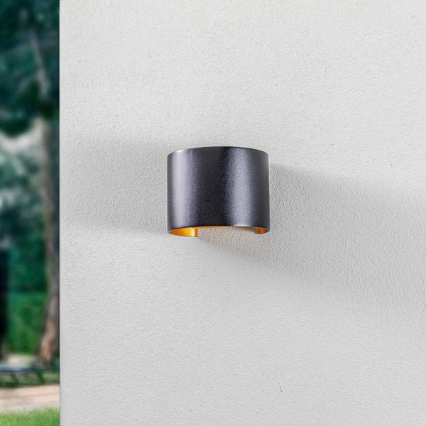 Zylindrische Wandleuchte in Schwarz mit goldener Innenseite, montiert an einer hellen Außenwand.