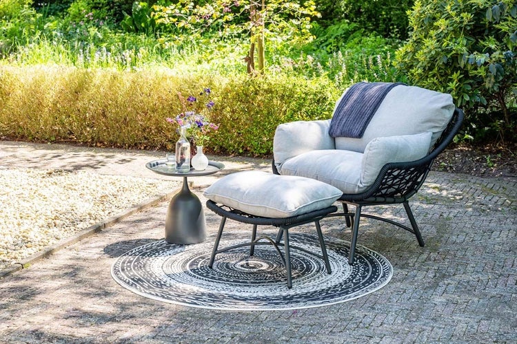 Moderner Garten-Lounge-Sessel mit Hocker, Beistelltisch und rundem Outdoor-Teppich auf einer gepflasterten Terrasse in einem hellen Garten.