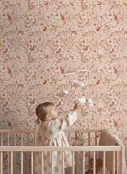 Kinderzimmer mit Kind im Holzgitterbett, das nach einem Mobile greift, vor einer Tapete mit Blumen- und Waldtiermuster.