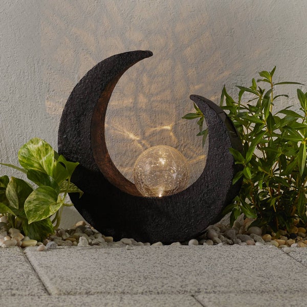 Solarleuchte in Form eines Sichelmonds mit leuchtender Glaskugel platziert zwischen Gartenpflanzen.