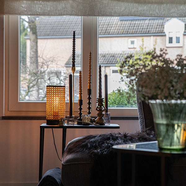 Dekorative Szene mit Tischlampe, Kerzen und Beistelltisch vor einem Fenster.