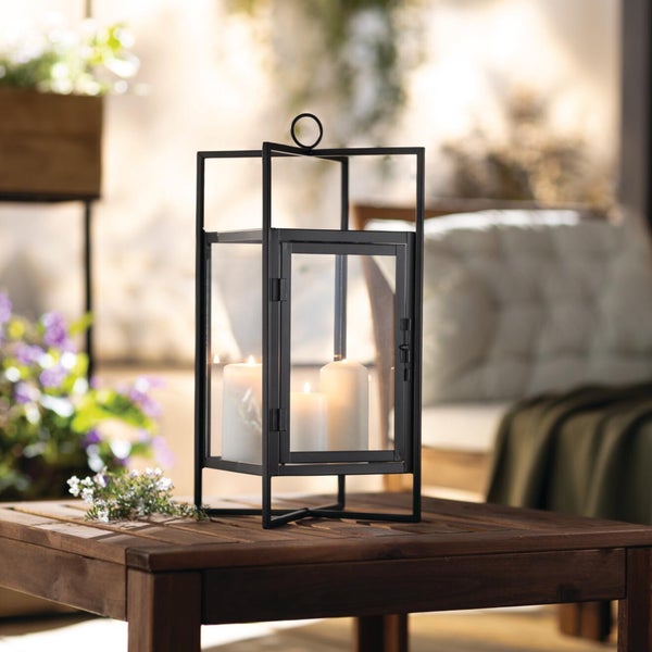 Moderne schwarze Metalllaterne mit Glasfenstern und drei brennenden Stumpenkerzen auf einem Holztisch im Freien.