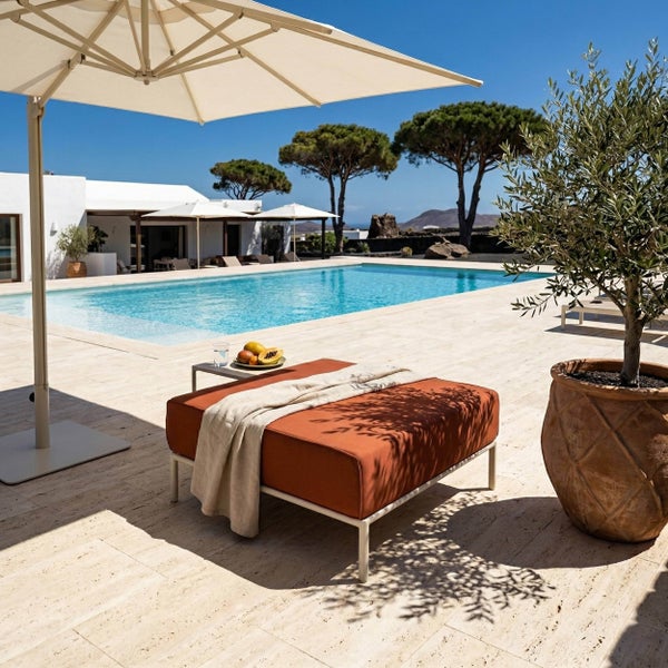 Moderner Gartenhocker mit rostfarbenem Kissen und Decke an einem Pool, neben einem Olivenbaum im Kübel und unter einem Sonnenschirm.