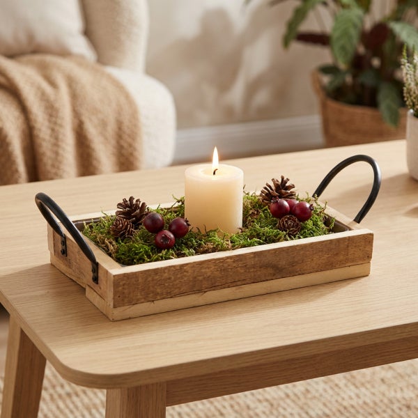 Dekoratives Holztablett mit Kerze, Zapfen und Moos als Tischdekoration auf einem Holztisch in einem Wohnraum.