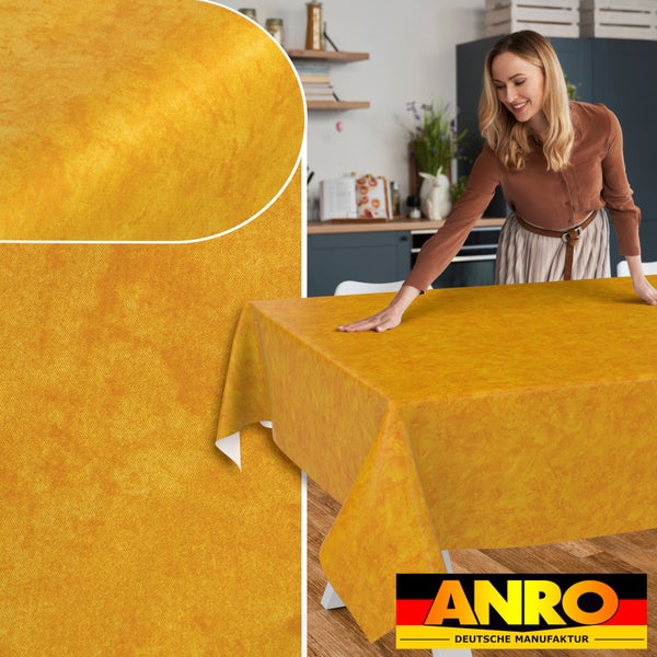 Frau glättet eine strukturierte Tischdecke auf einem Tisch in einer hellen, modernen Küche. Detailansicht des Musters. ANRO DEUTSCHE MANUFAKTUR Logo.