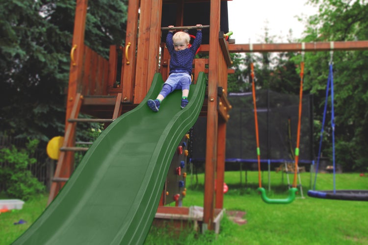 Kind sitzt oben auf einer grünen Wellenrutsche an einem Spielturm aus Holz im Garten.