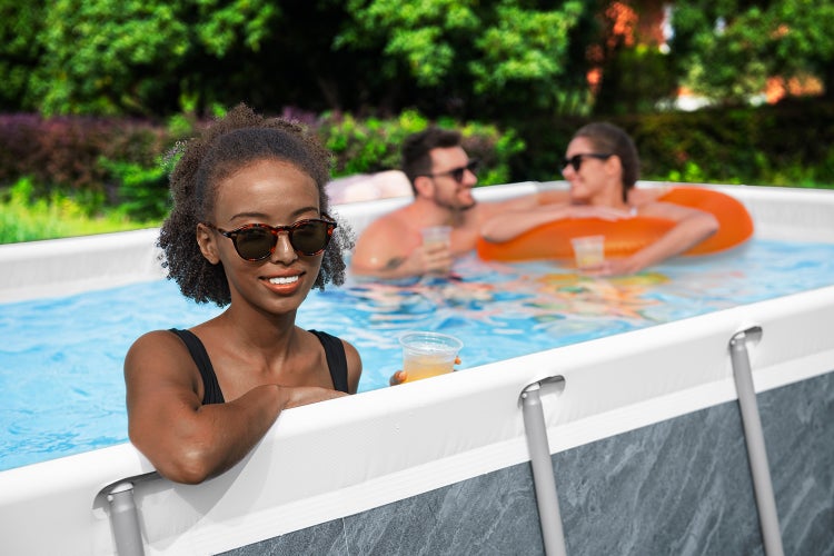Drei Personen entspannen in einem rechteckigen Aufstellpool mit Steinoptik-Außenwand im Garten.