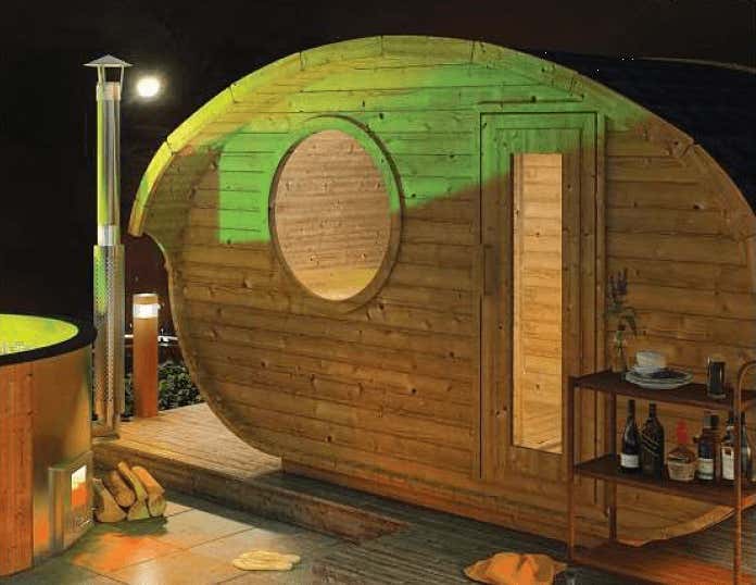Ovale Gartensauna aus Holz in Fassform mit rundem Fenster und Glastür auf einer Terrasse neben einem Badefass bei Nacht.