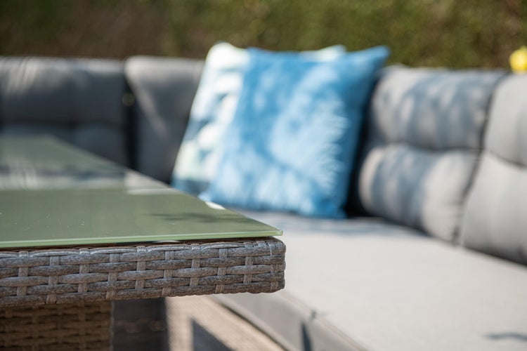 Nahaufnahme eines Gartentisches mit Glasplatte und grauem Kunststoffgeflecht neben einem Loungesofa mit grauen und blauen Kissen.