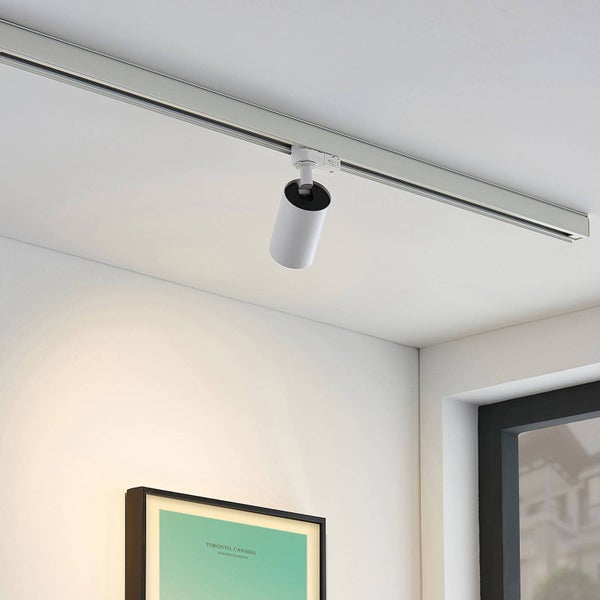 Moderner weißer Schienenstrahler an einer Deckenschiene in einem hellen, minimalistischen Raum mit Bilderrahmen und Fenster.