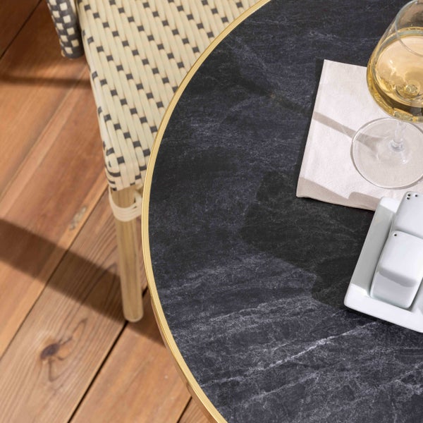 Runder Bistrotisch mit Tischplatte in dunkler Marmoroptik und goldfarbenem Rand auf einer Holzterrasse neben einem Flechtstuhl.
