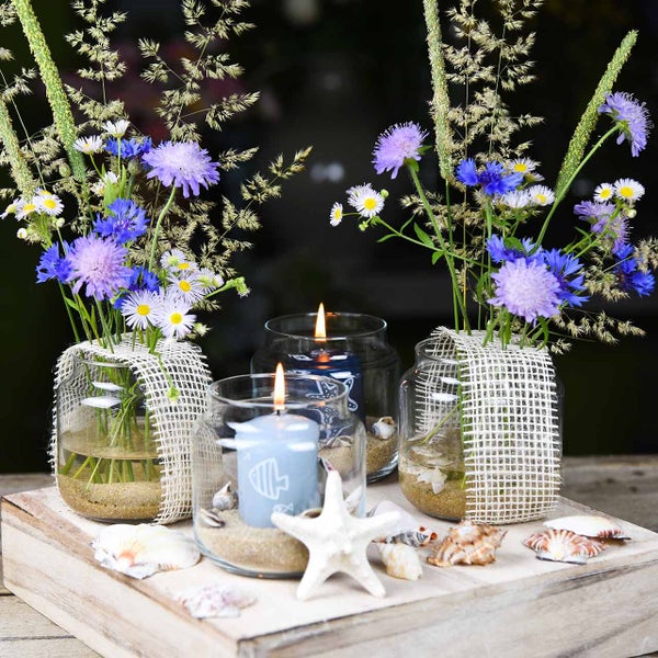 Dekorative Tischdekoration mit Blumen, Kerzen und Muscheln in Gläsern auf einem Holztablett