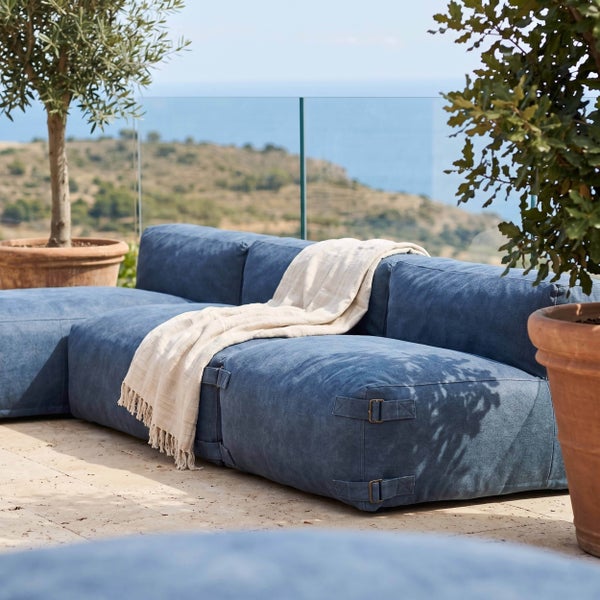 Modernes blaues Outdoor-Loungesofa auf einer Terrasse mit Glasgeländer, Decke und Terrakotta-Pflanzkübeln vor einer Küstenlandschaft.
