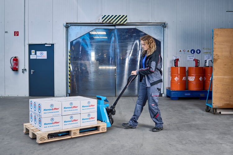 Frau in grauer Arbeitskleidung bedient blauen Hubwagen mit beladener Holzpalette in einer hellen, organisierten Lagerhalle.