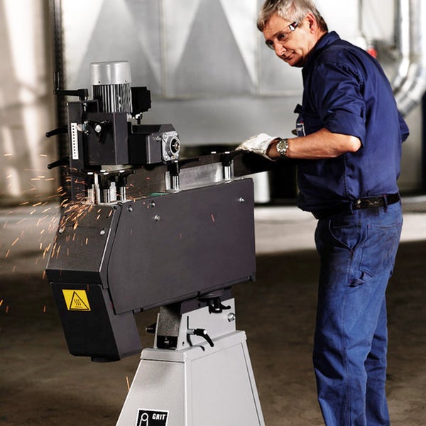 Ein Mann mit Schutzbrille bedient eine GRIT Bandschleifmaschine zur Metallbearbeitung in einer Werkstatt unter Funkenflug.