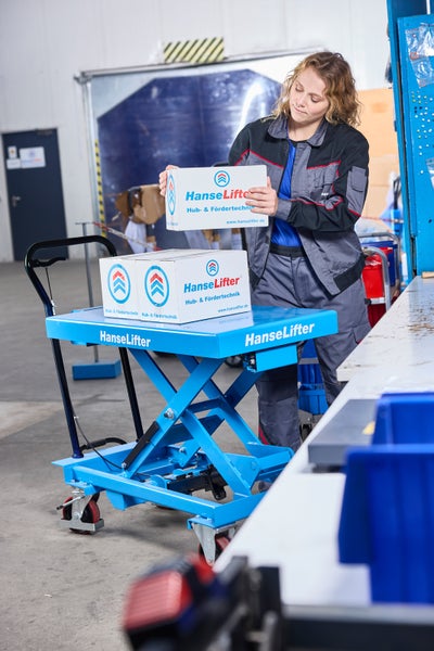 Frau in Arbeitskleidung belädt in einer hellen Werkstatt einen blauen HanseLifter Scherenhubtisch mit weißen Kartons.