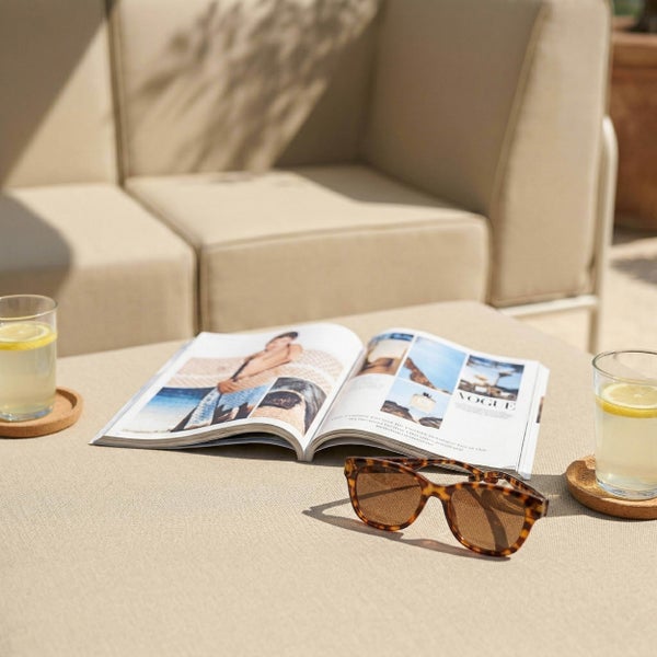Helle Garten-Lounge mit beigen Polstermöbeln, aufgeschlagener Zeitschrift, Sonnenbrille und Getränken auf einem Tisch.