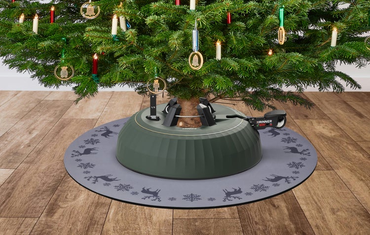 Krinner Christbaumständer mit Fußhebel auf einer grauen Unterlage mit Rentier-Motiven unter einem geschmückten Weihnachtsbaum auf Holzboden.