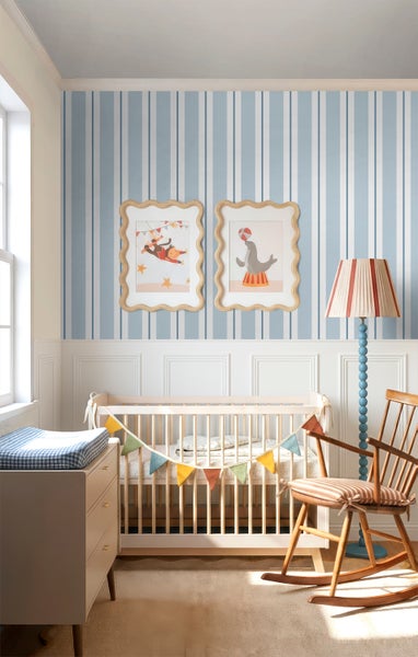 Helles Kinderzimmer mit blau-weiß gestreifter Tapete, weißer Wandtäfelung, Babybett aus Holz, Schaukelstuhl und Wandbildern mit Zirkusmotiven.