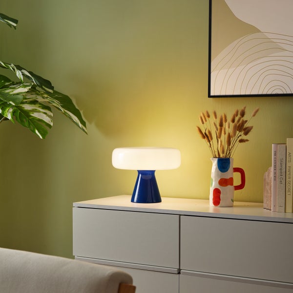 Moderne pilzförmige Tischleuchte mit blauem Fuß und weißem Schirm auf einer weißen Kommode in einem hellen Raum mit dekorativen Elementen.