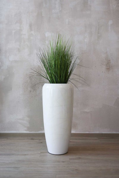 Künstliches Gras in einer hohen weißen Bodenvase vor einer Wand in Betonoptik.