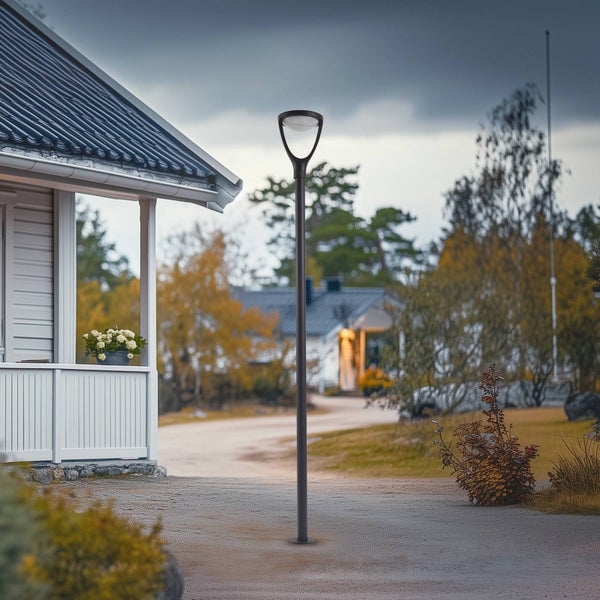 Moderne Mastleuchte in Schwarz vor einem Haus mit weißer Holzveranda, schlankes Design mit dreieckigem Leuchtenkopf.