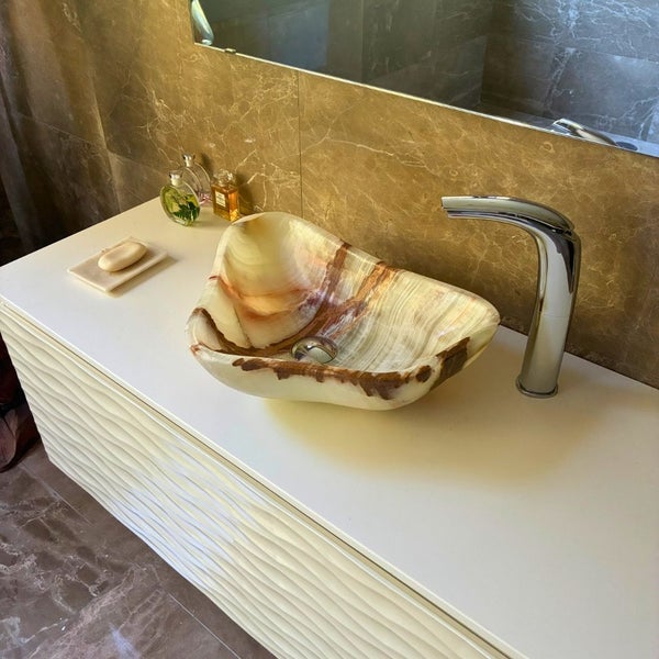 Aufsatzwaschbecken aus Naturstein in organischer Form auf einem weißen Waschtisch mit gewellter Front und verchromter Armatur.
