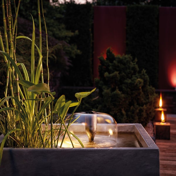Moderner Gartenbrunnen in einem grauen Pflanzkübel mit LED-Beleuchtung und Wasserglocke, umgeben von Gräsern auf einer Terrasse am Abend.