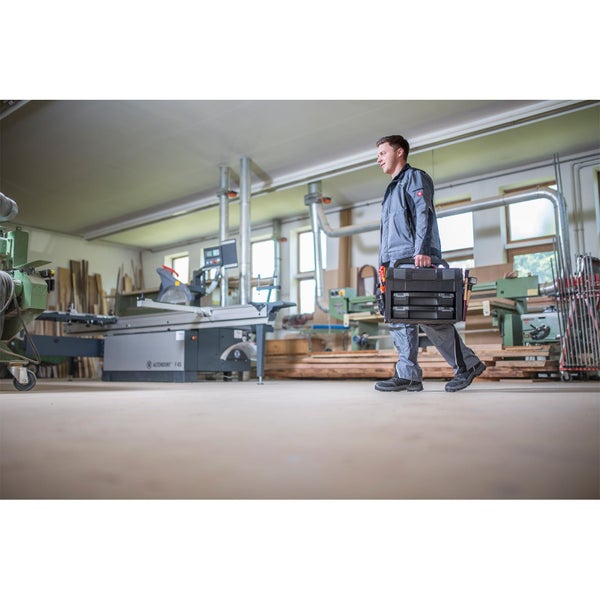 Handwerker trägt einen schwarzen Werkzeugkoffer mit Schubladen durch eine helle, geräumige Holzwerkstatt mit einer großen Formatkreissäge.