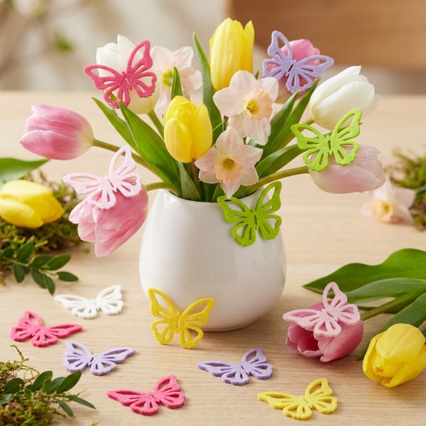 Osterstrauß aus Tulpen und Narzissen in einer weißen Vase, dekoriert mit bunten Filzschmetterlingen als Tischdekoration.