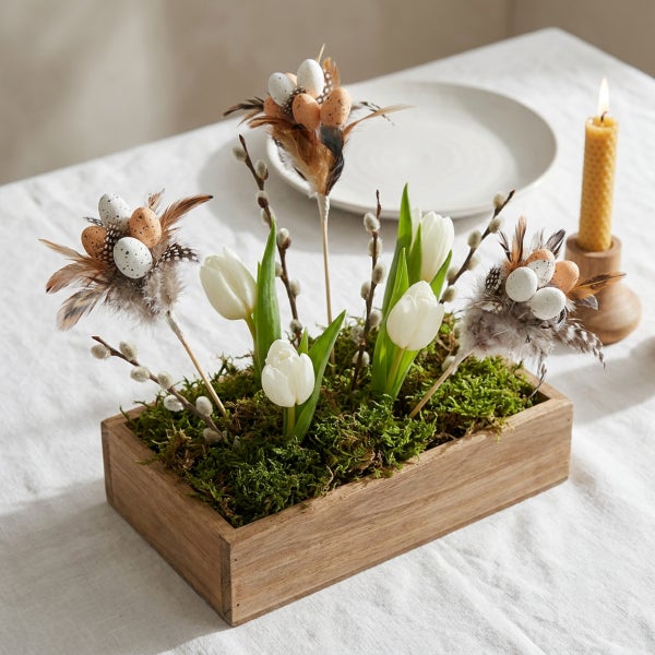 Ostergesteck im Holzgefäß mit weißen Tulpen Weidenkätzchen Federn und kleinen Ostereiern auf einem Tisch arrangiert