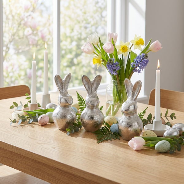 Osterdekoration mit drei silbernen Hasenfiguren, einer Blumenvase mit Frühlingsblumen, Stabkerzen und Ostereiern auf einem Holztisch.