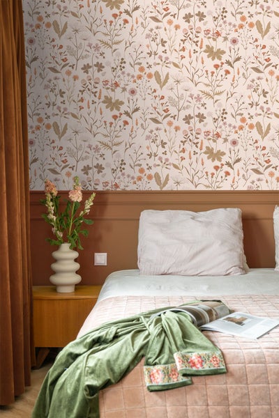 Vliestapete mit floralem Wiesenblumen-Muster in einem Schlafzimmer über einer braunen Wandpaneele neben einem Bett mit Textilien.
