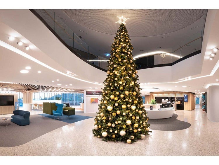 Großer künstlicher Weihnachtsbaum mit festlicher Beleuchtung und Kugelschmuck in einem modernen, offenen Bürogebäude mit Empfangsbereich.