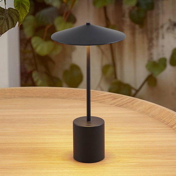 Schwarze, moderne Tischleuchte mit schmalem Standfuß und flachem Schirm auf einem Holztisch vor Pflanzenhintergrund.