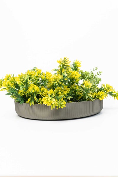 Künstliche Pflanze mit gelben Blüten in einer flachen, grauen, gerippten Schale.