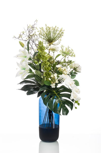 Künstliches Blumengesteck mit weißen Lilien, Orchideen und einem Monstera-Blatt in einer blauen Glasvase.