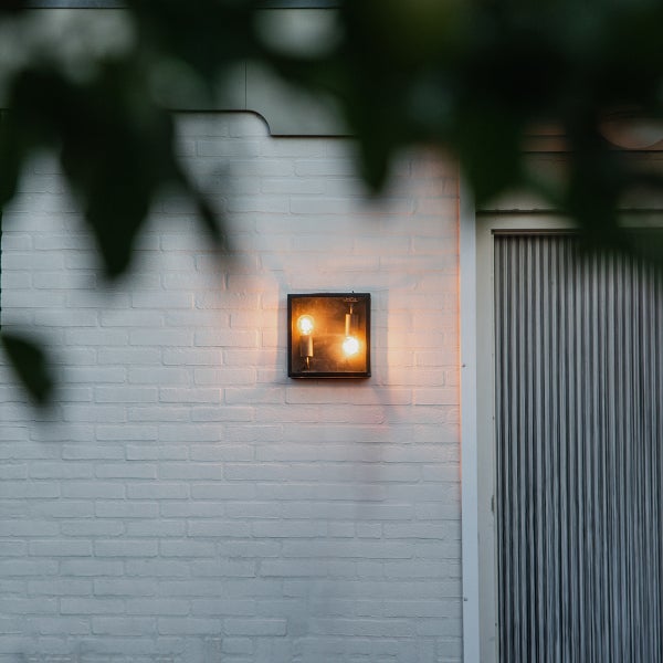 Moderne Außenwandleuchte mit schwarzem Rahmen und zwei leuchtenden Leuchtmitteln an einer weißen Ziegelwand neben einer Tür.