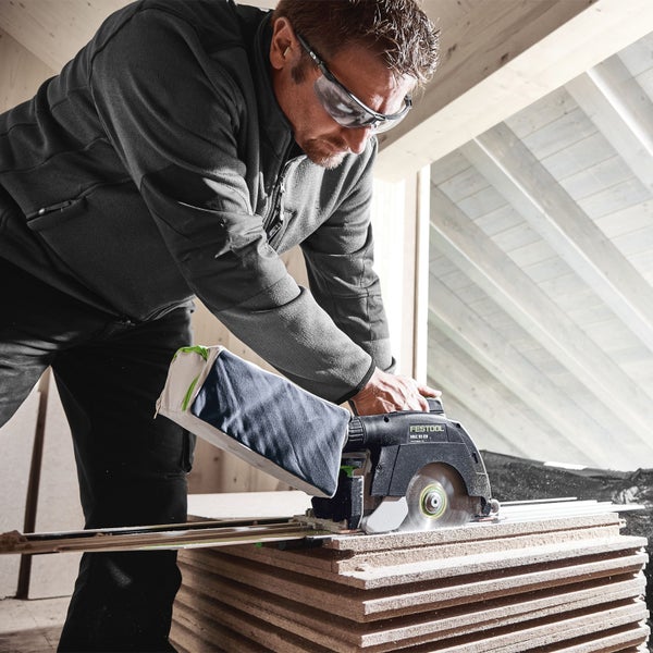 Ein Mann sägt mit einer Handkreissäge Holzbretter. Er trägt eine Schutzbrille. An der Säge ist ein Staubfangsack befestigt.