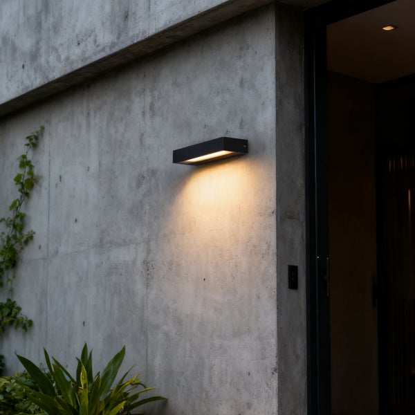 Moderne rechteckige Außenwandleuchte in Schwarz an einer Sichtbetonwand montiert, nach unten gerichteter Lichtkegel neben einem Eingangsbereich.