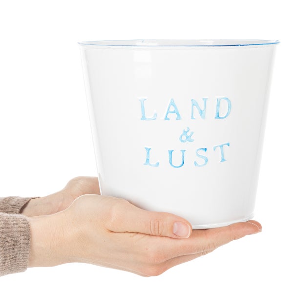 Weißer Metallübertopf mit Prägeschrift LAND und LUST, in Händen gehalten.