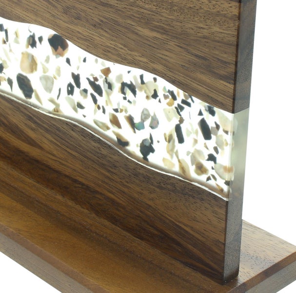 Nahaufnahme eines Holzpaneels mit einer wellenfoermigen Einlage aus lichtdurchlaessigem Harz mit kleinen Dekosteinen in verschiedenen Nuancen.